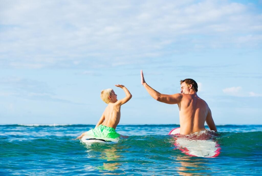 Father and son beginner surfers high-fiving on their surfboards during a family surf lesson in the Azores
