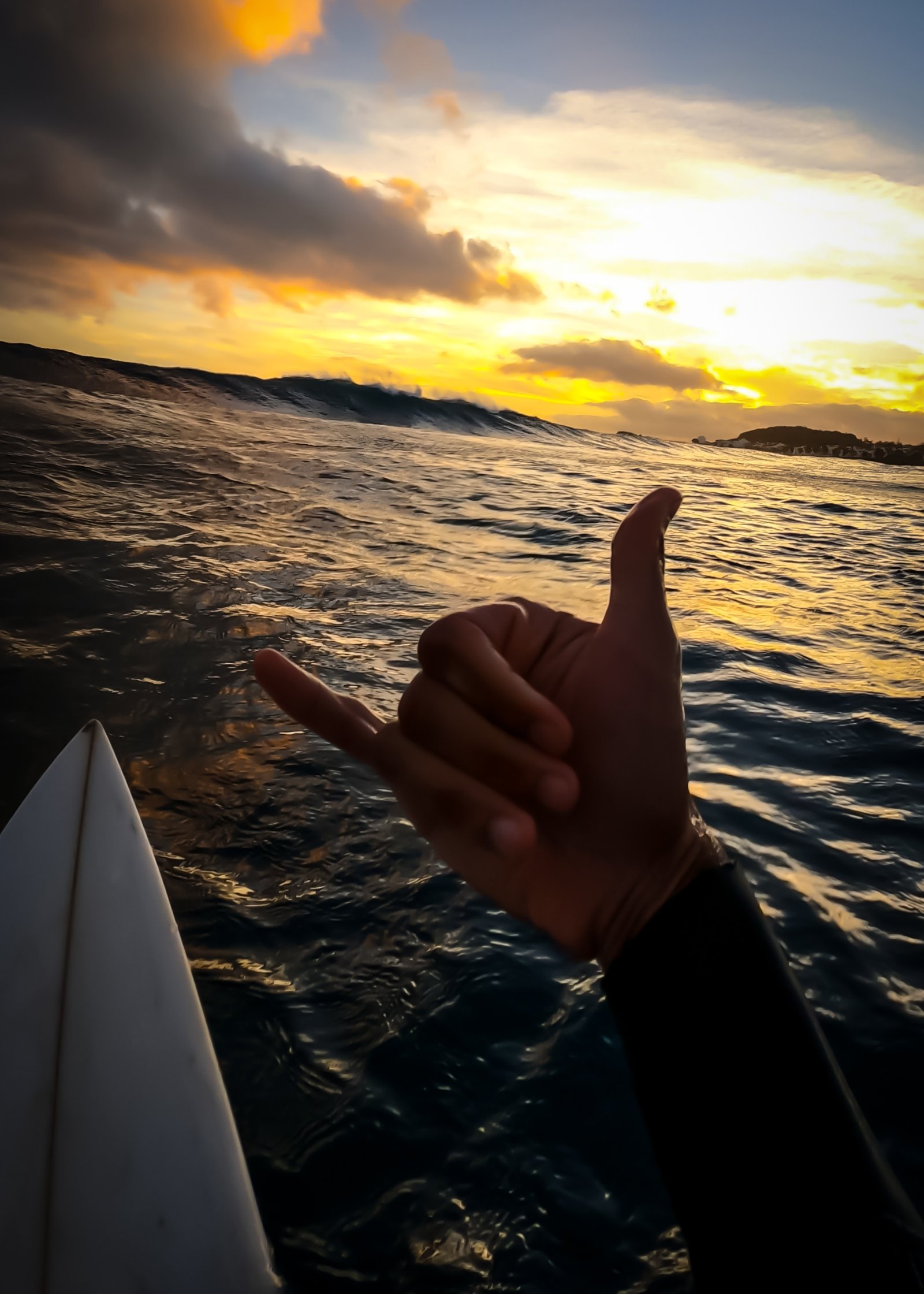 View from the water showing a breaking wave, surfboard nose, and a shaka at sunset