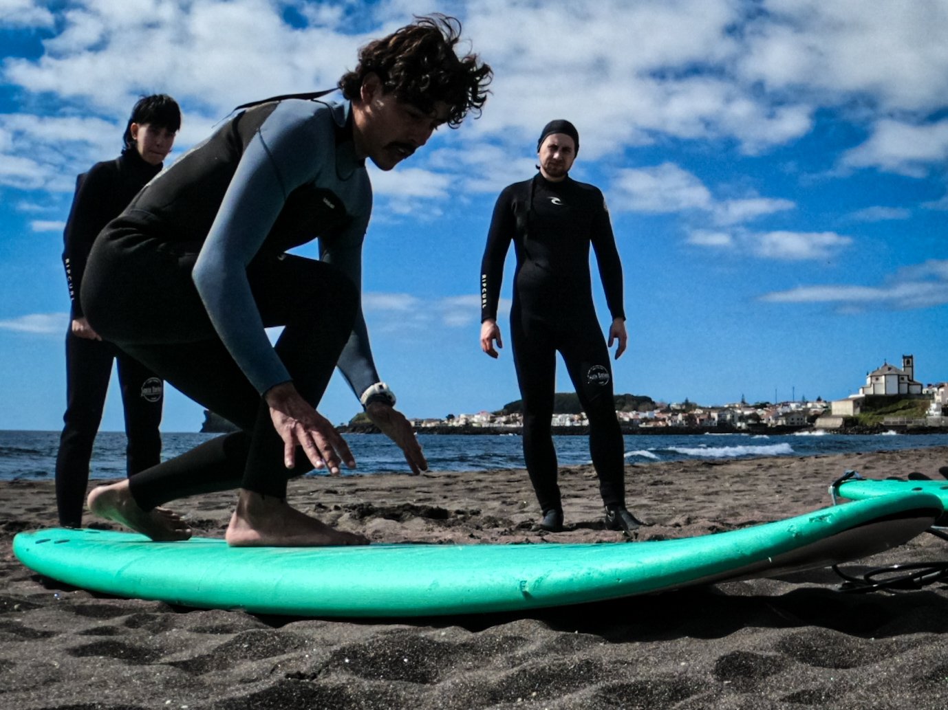 A surf coach teaching two beginners on the beach how to stand up on the soft top surfboard.